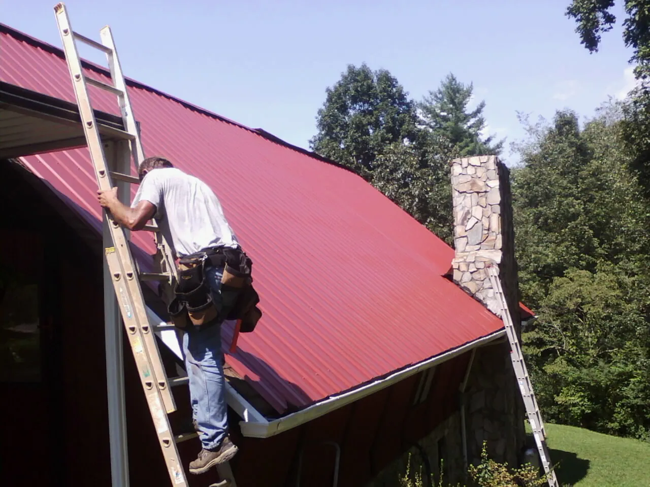 Roofer on a ladder beside a red metal roof and stone chimney — Soffit & Fascia Repair by Roofing Brothers in Zimmerman